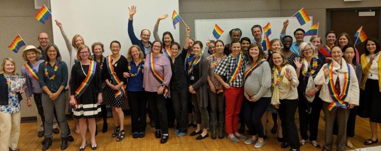 A group of people line up and smile while waving rainbow flags